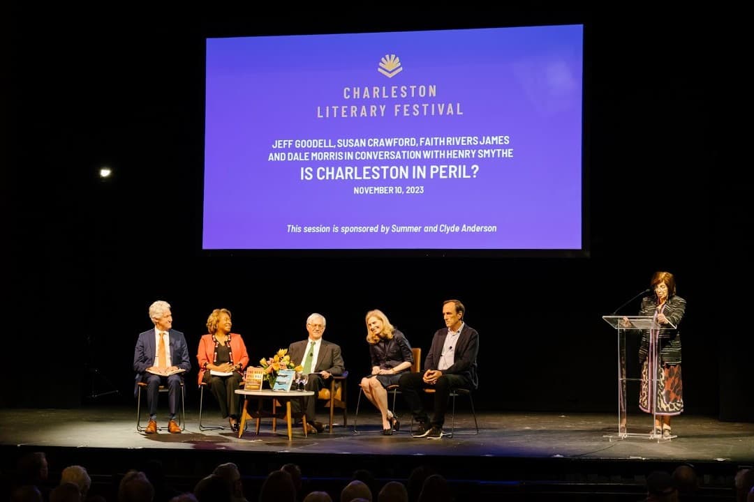Five panelists seated on a lit stage below a large screen reading 'Charleston Literary Festival, Jeff Goodell, Susan Crawford, Faith Rivers James and Dale Morris in conversation with Henry Smythe, Is Charleston in Peril?, November 10, 2023', with a woman standing at a podium to the right addressing the audience